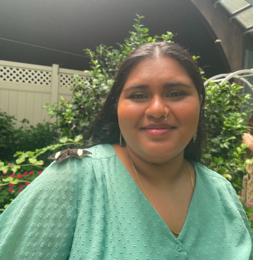 Pavi Parthiban in front of green foliage and a white fence at night. Pavi is wearing a teal blue blouse with a button and has on a gold chain, a golden nose piercing, and golden earrings. Pavi's dark hair is thrown behind.