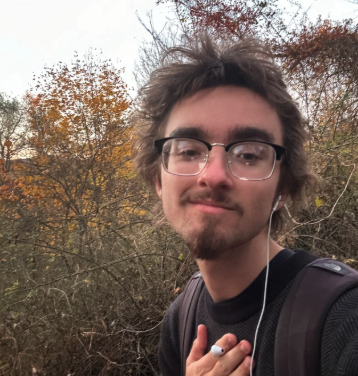 Aiden O'Neill in front of brown foliage during the day. Aidan is wearing a black t-shirt with a black backpack and black and silver glasses and has one white wired earbud in ear and is holding the other. Aidan has brown hair with a goatee/mustache