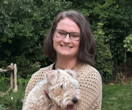 Annie Riley is outdoors in front of foliage holding a white scotty dog. The dog is wearing a red collar and is panting. Annie is wearing a tan mesh-like shirt with black square glasses and has shoulder length brown hair