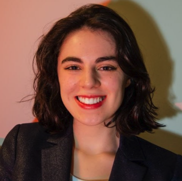 Marina Goldman smiling direct to camera wearing a black suit coat. Marina's hair is just above the shoulders and dark and wavy. Marina has on red lipstick