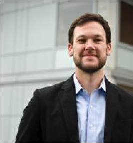 Thaddeus Miller in front of a white building wearing a black suit with a light-blue button up. Thaddeus has dark hair and a short beard.