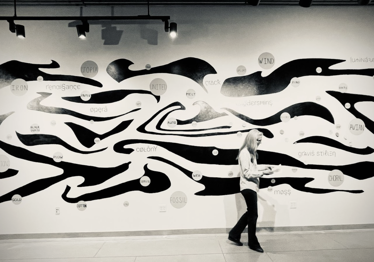 A woman looks to the right of the image and down, holding a piece of paper in front of a wall with black squiggles on it interspaced with circles. Some of the circles have visible words like Wind, Fossil, Iron, Utopia, Melt, colony, Plow, and Gold