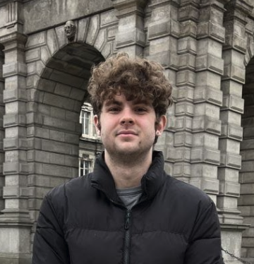 Eric Strong stands in front of a stone archway. Eric is wearing a black puffer jacket with a grey t-shirt poking out. Eric has curly brown hair and a small smile