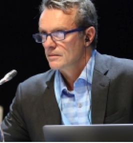 Rob Deconto presenting in front of a small microphone and laptop wearing a grey suit with a blue and white checkered collared shirt. Rob has on black glasses and is wearing one wired headphone