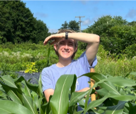 Grace Ziegel is standing behind a short row of green foliage and in front of a nature background. Grace's left arm covers the face from sun and has on a black watch. Grace is wearing a light blue tshirt.