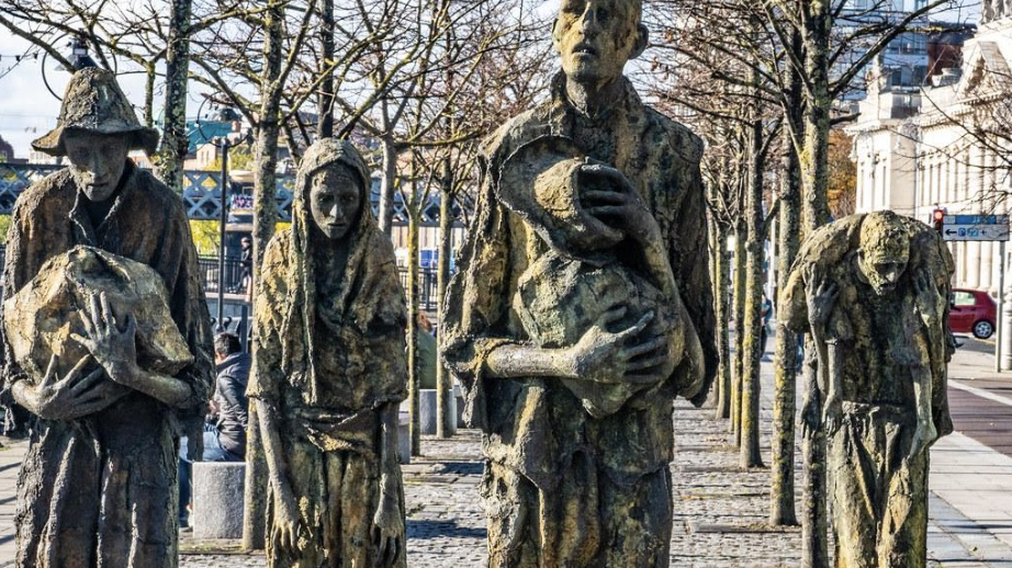 Irish Famine Memorial in Dublin showcasing four figures cast in metal that has decayed. The figures are gaunt with haunted faces. Some are carrying bundles and the man on the far right has a limp child slung over his shoulders