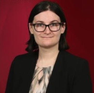 Kylie Bane looking straight to camera wearing a brown and white geometric blouse under a black blazer with black square glasses.  Kylie's hair is dark and cut just above the shoulder