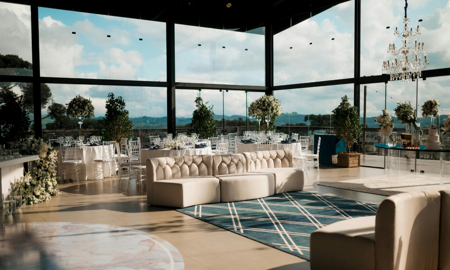 Elegant event space with large windows, white sofas, round tables with floral centerpieces, a chandelier, and a view of the outdoors with blue sky and clouds.