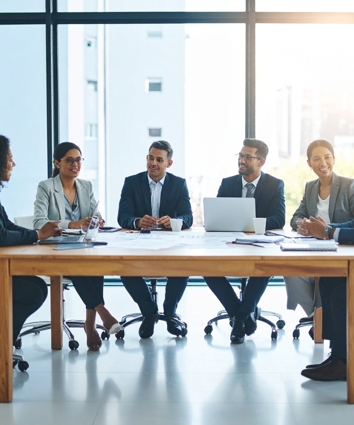 Six business professionals sitting around a wooden conference table in a modern office, engaged in a discussion, with large windows and a cityscape in the background.