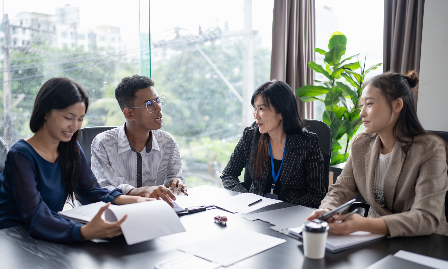 Four diverse women in business attire are having a meeting in a modern office with large windows and a green plant, discussing documents and using a smartphone.