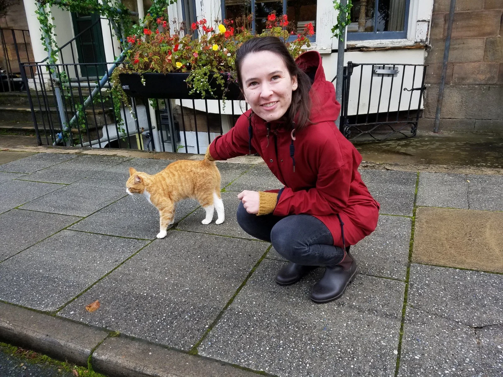 Wearing a red raincoat, Joan squats on a sidewalk in Harrogate (UK) while petting an orange tabby cat and smiling at the camera. At the time of this photo, Joan was studying Medieval Latin, Old Norse, and Old English at the University of York.
