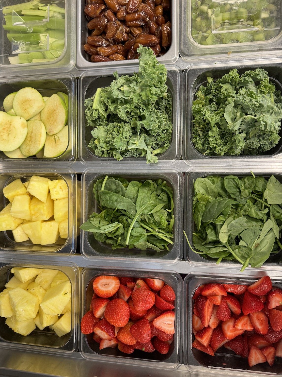 Plastic containers filled with sliced strawberries, pineapples, apples, spinach, kale, celery, and green grapes.