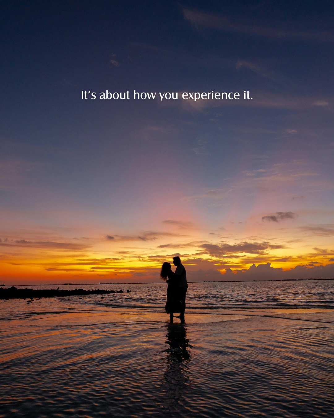 Couple standing in shallow water at sunset on the beach, with the text 'It's about how you experience it' overlaid.