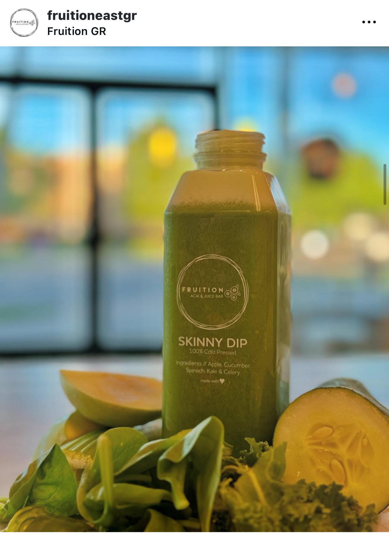 A green bottle labeled 'Skinny Dip' with ingredients listed as apple, cucumber, spinach, kale, and celery, surrounded by sliced apple and green vegetables, with a blurred cityscape background visible through a window.