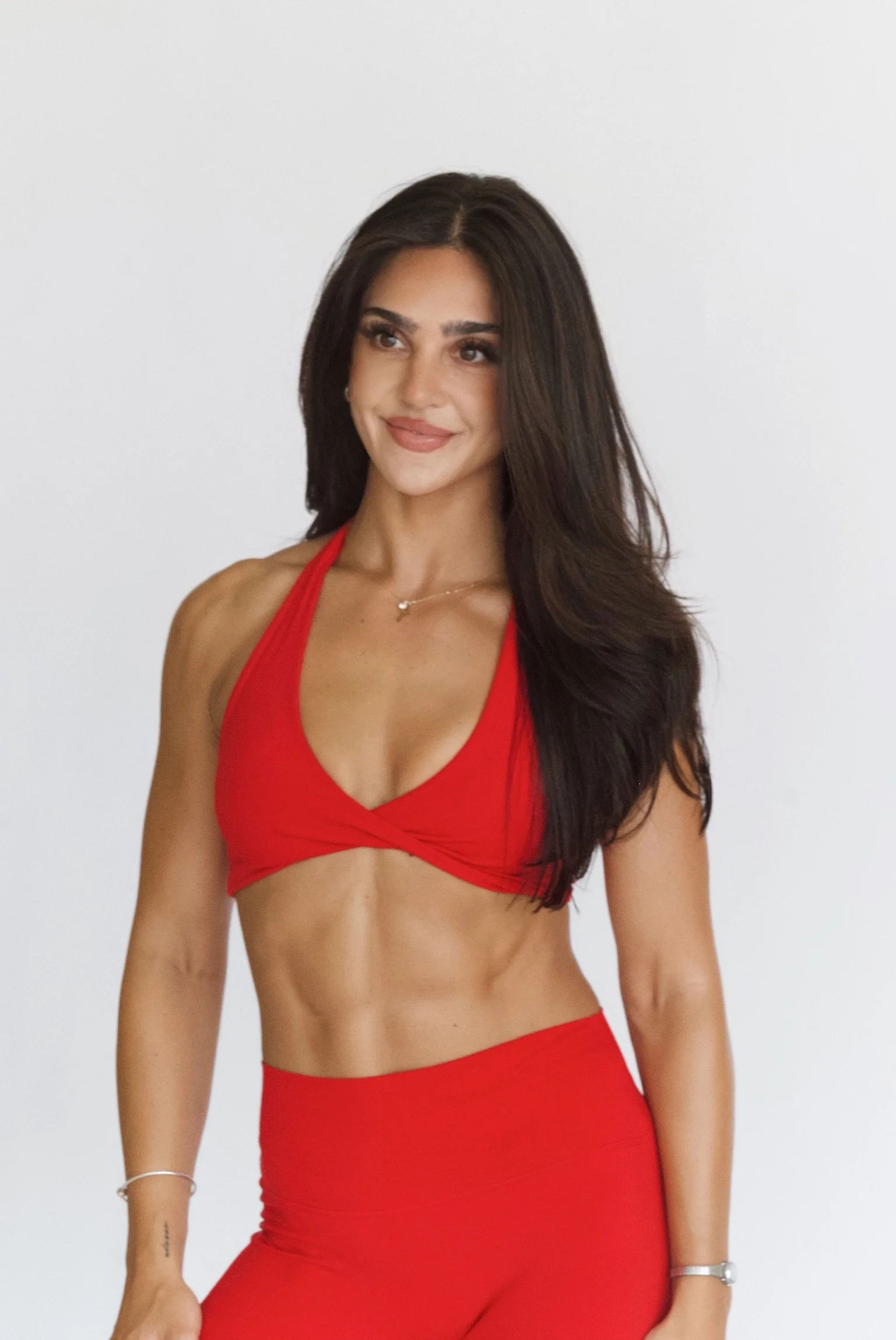 A woman with long dark hair wearing a red sports bra and matching red leggings, smiling, standing against a plain white background.
