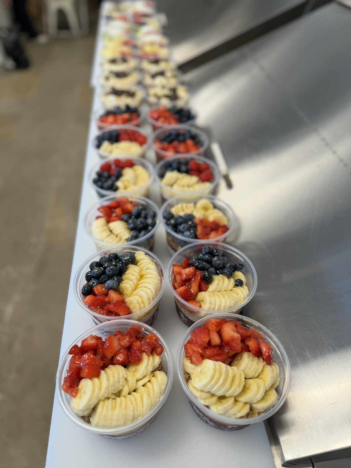 Plastic cups filled with layered dessert topped with sliced bananas, strawberries, and blueberries, arranged on a long white table.