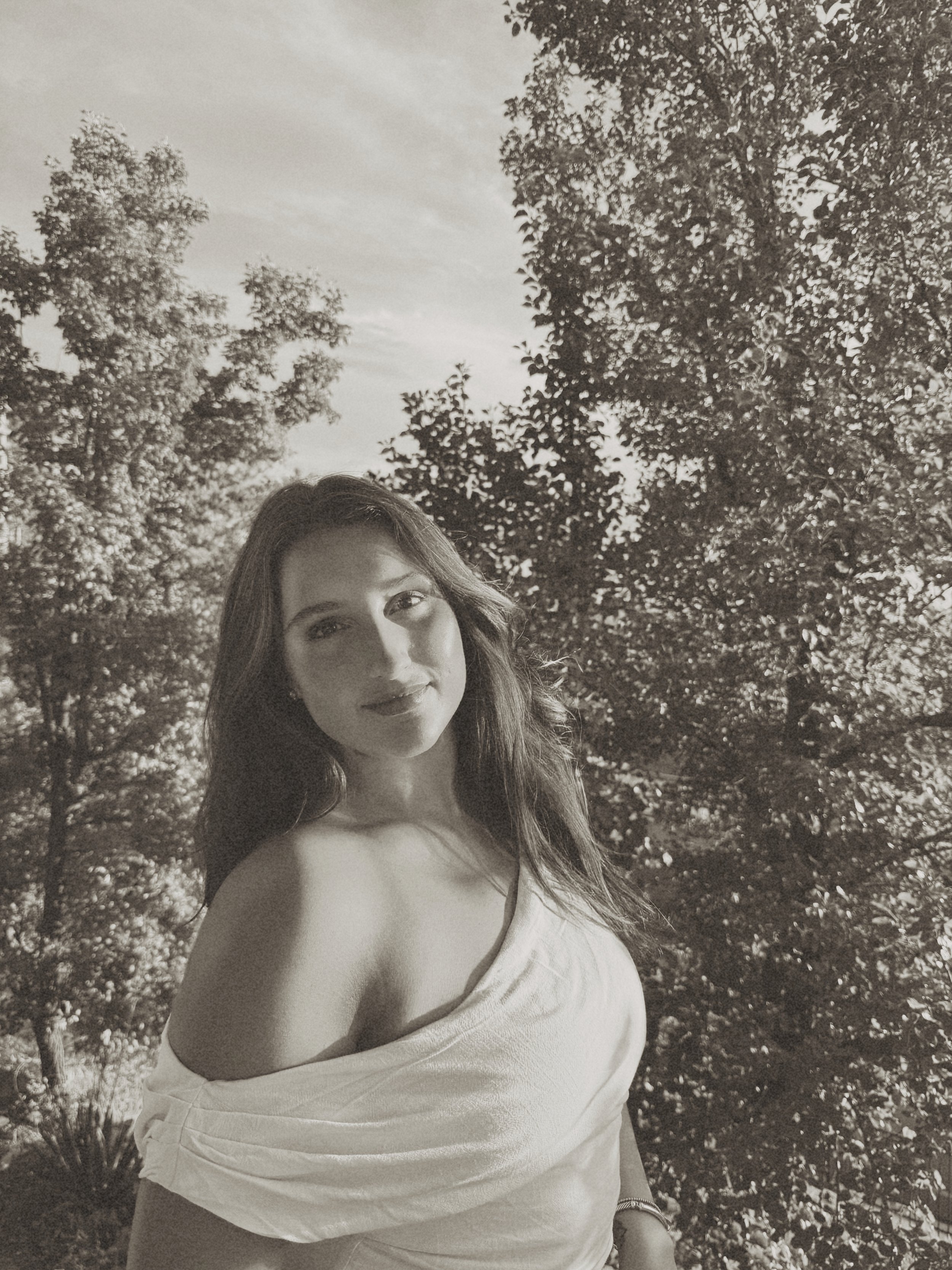 A young woman with long hair standing outdoors in front of trees, wearing a white off-the-shoulder top, looking at the camera, in black and white.