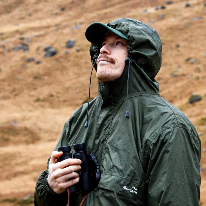 A man dressed in a green waterproof jacket with a hood and cap, holding binoculars, standing outdoors on a hillside with a rocky terrain.