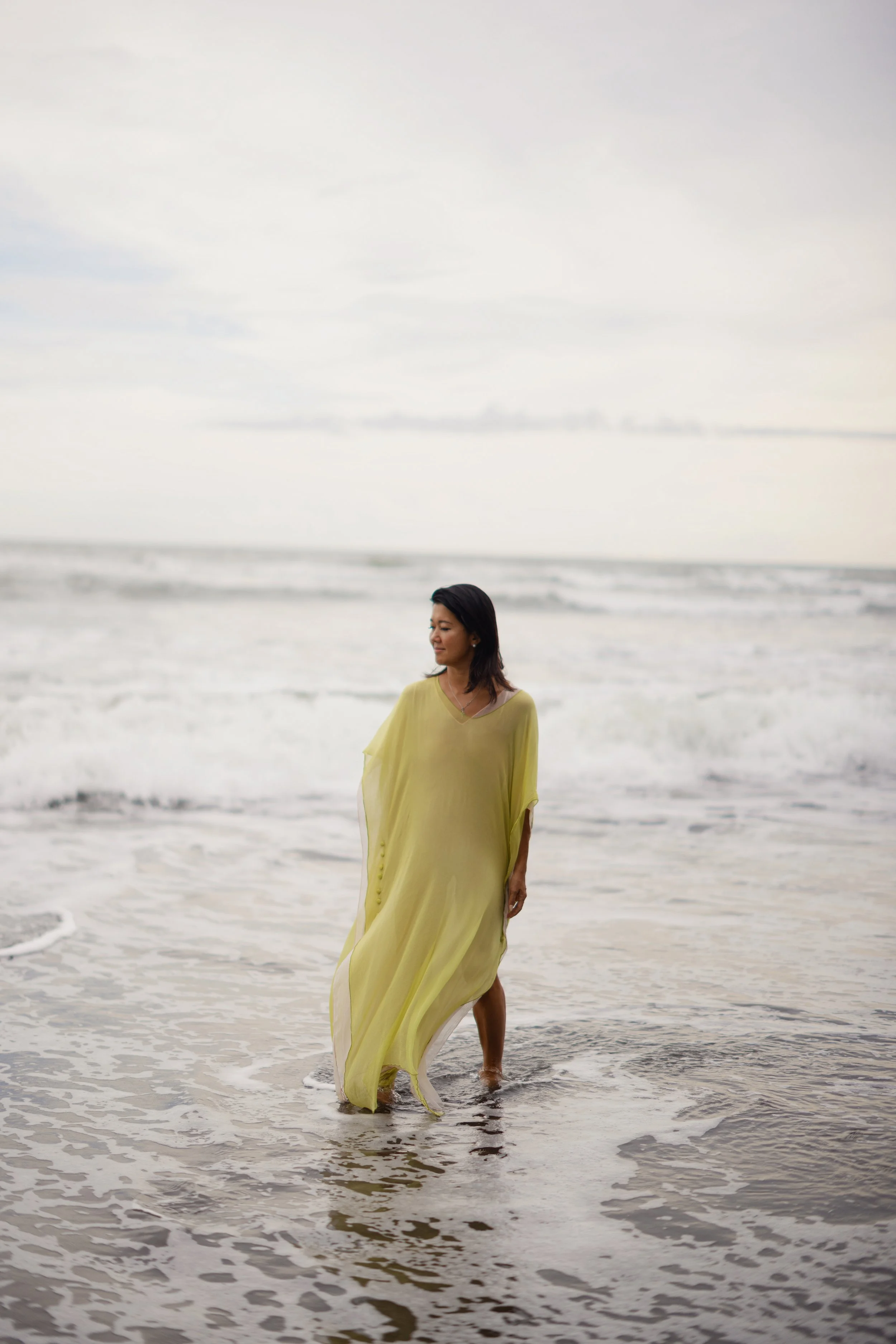 A woman in a yellow dress walking in the ocean water at the beach, with an overcast sky in the background.