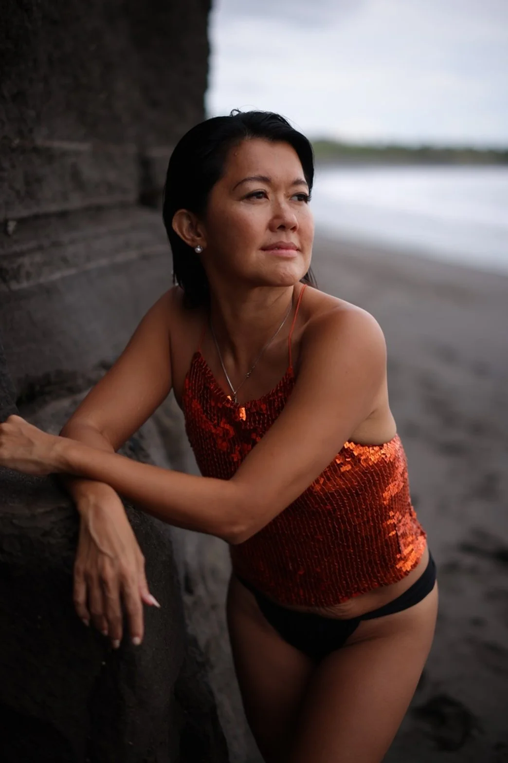 Woman with short dark hair wearing a red sequin tank top and black bottoms, leaning against a rock by the beach, looking thoughtfully into the distance.