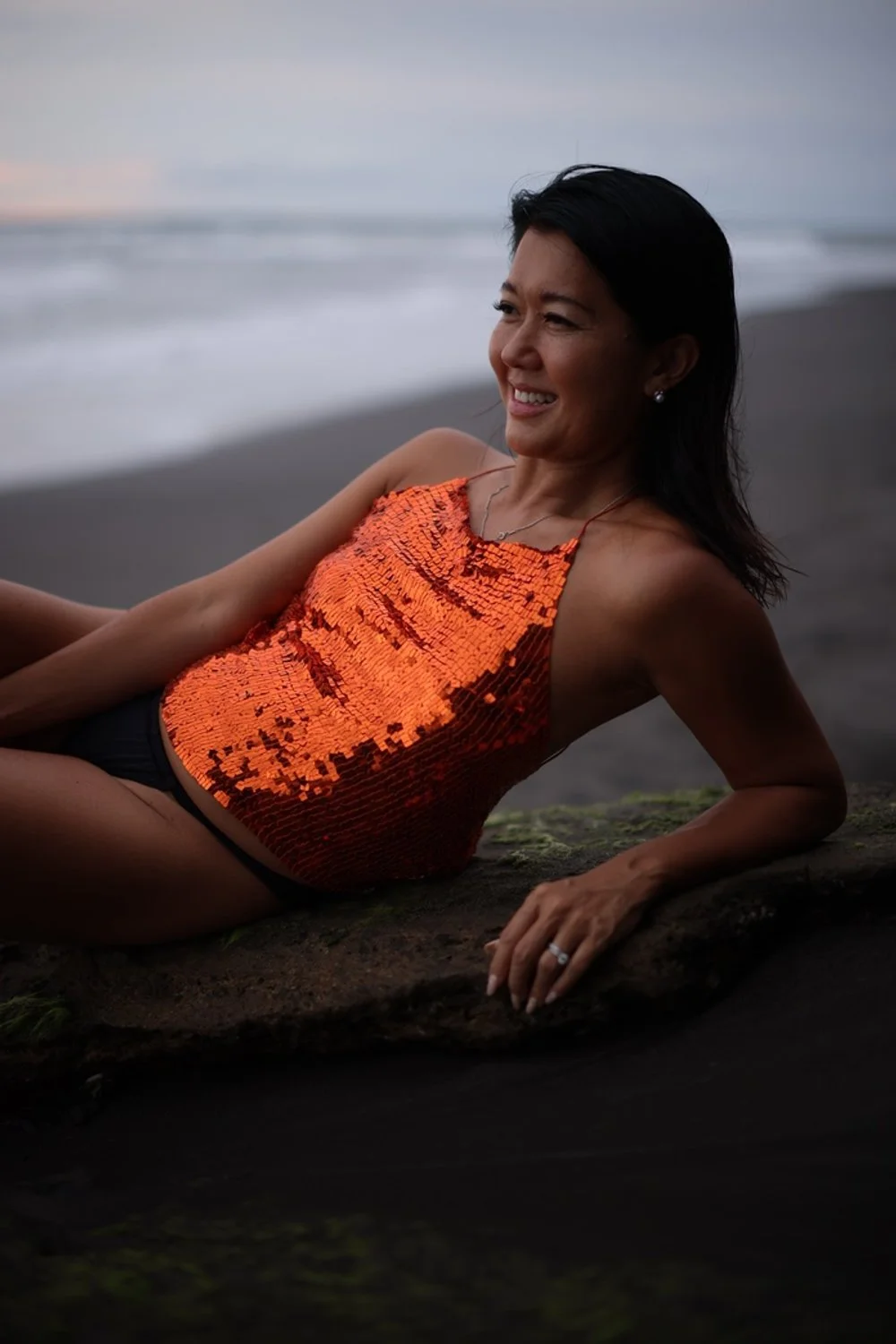 A woman lying on a rock at the beach during sunset, wearing an orange sequin top and dark bottoms, smiling.