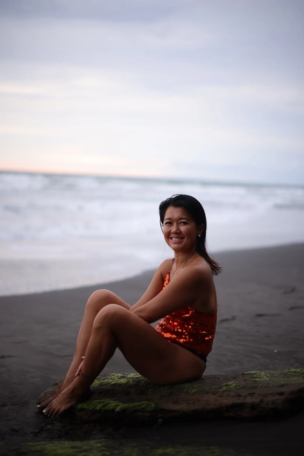 Woman in orange sequin swimsuit sitting on a moss-covered log on the beach with waves and an overcast sky in the background.