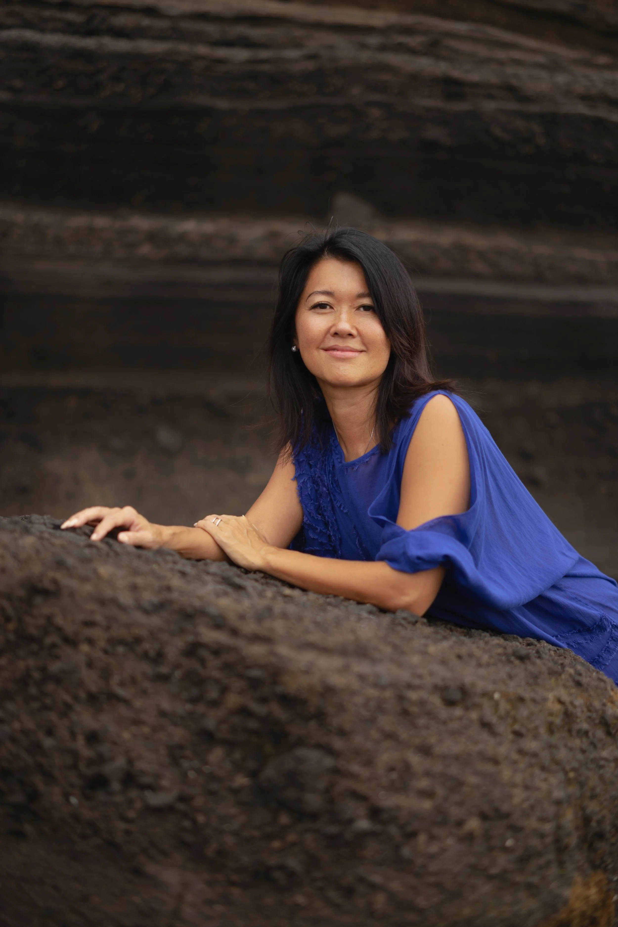 A woman in a blue dress leaning on a rock with a dark, rocky background.