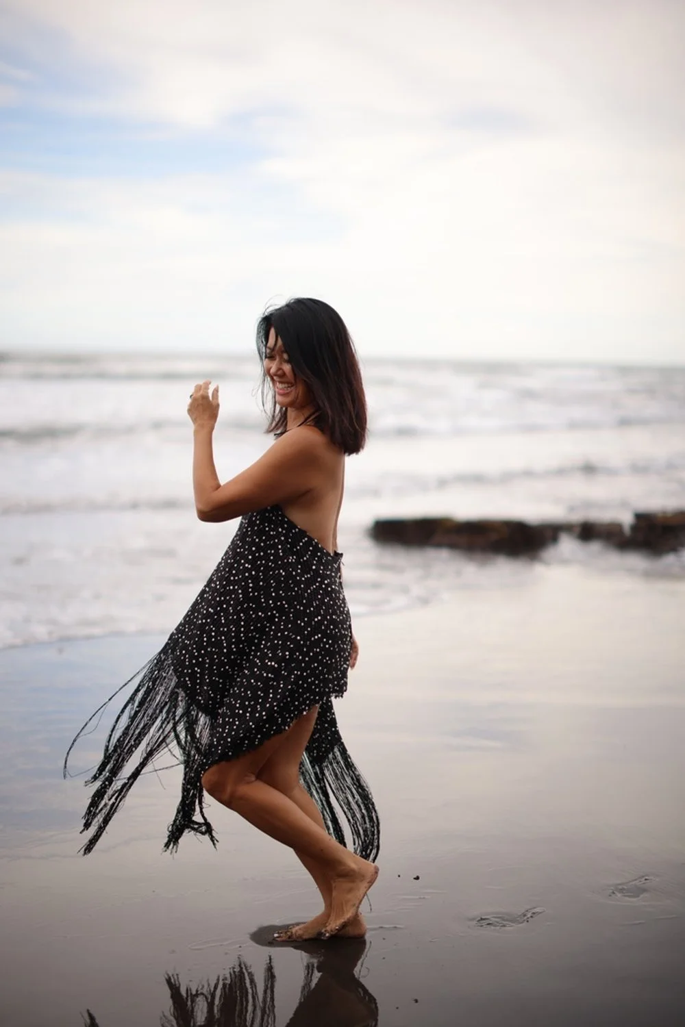 A woman smiling and laughing at the beach, standing on wet sand with waves in the background, wearing a black and white polka dot dress with fringes, barefoot.