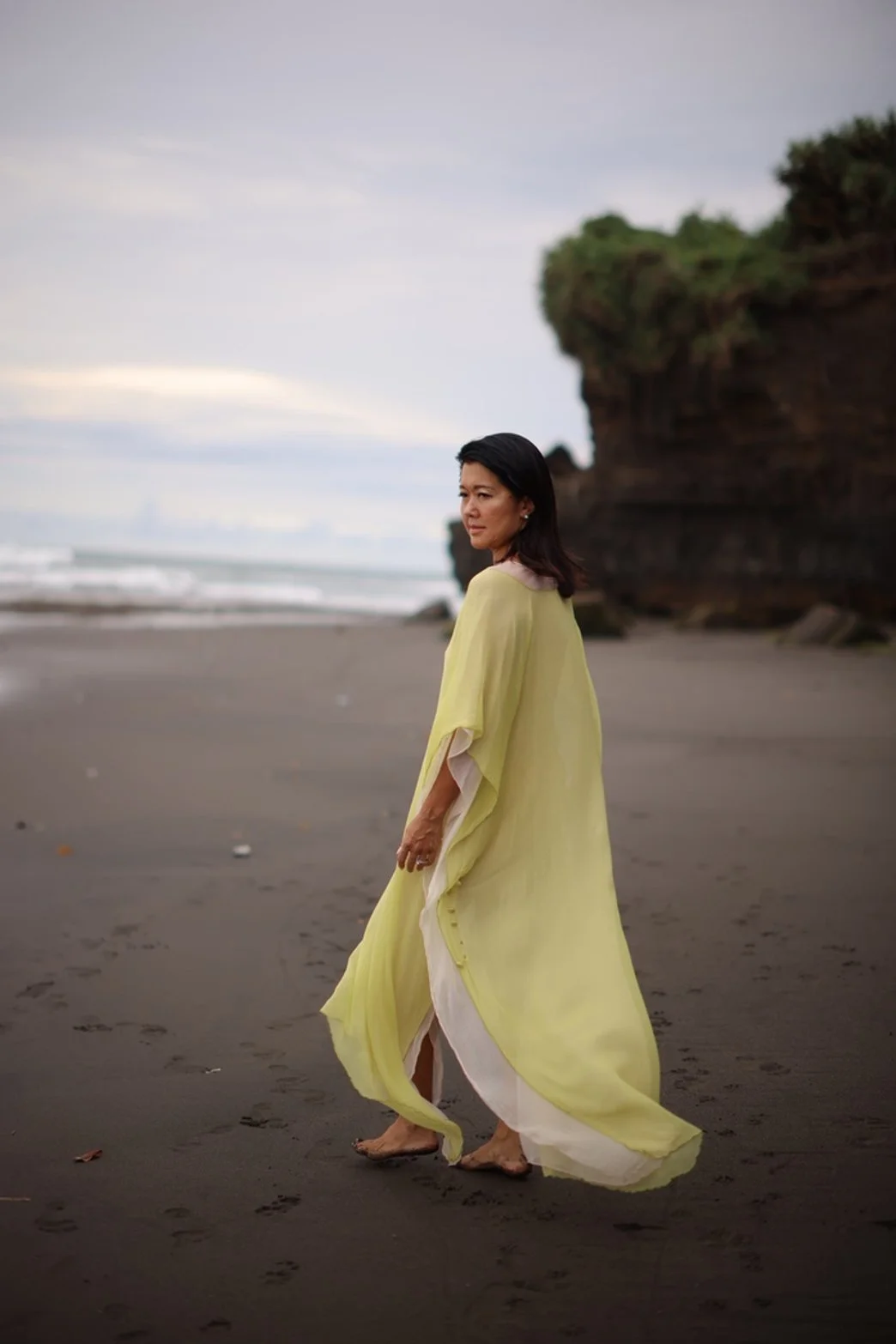 Woman in a flowing yellow dress walking barefoot on a dark sandy beach with rocky cliffs in the background.