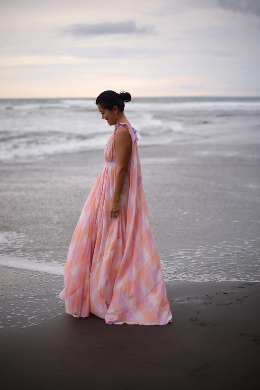 Woman in a flowing pink, orange, and white patterned dress standing on the beach near the shoreline with waves and a cloudy sky in the background.