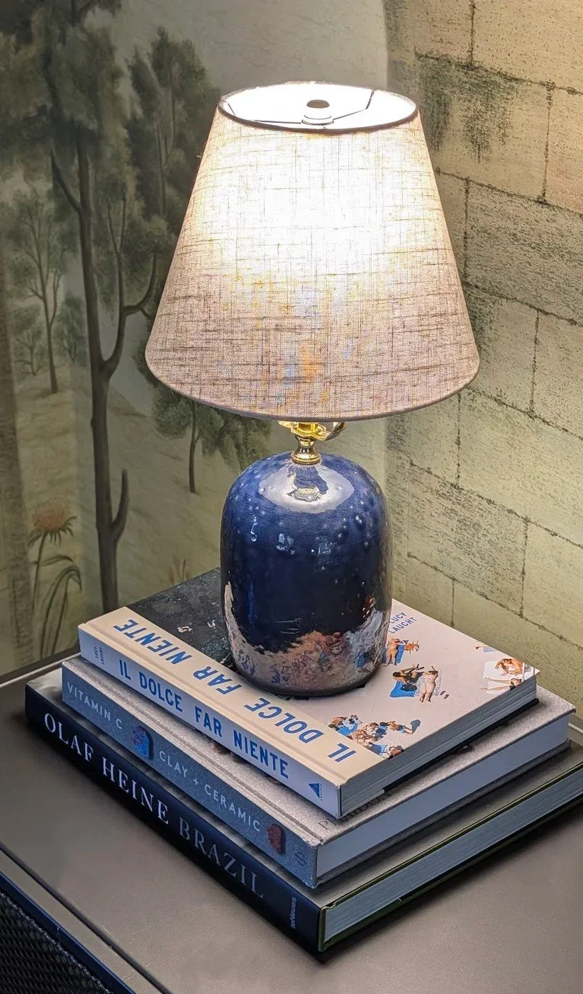 A table lamp with a blue ceramic base and beige fabric shade placed on top of four hardcover books on a table. In the background, there is a painted wall with a nature scene and an exposed brick wall.