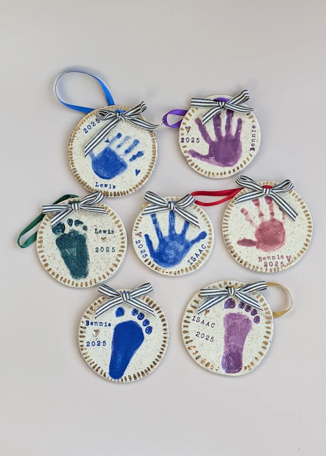 Set of seven circular handprint ornaments in various colors with black-and-white striped bows and ribbons, each engraved with a name, date, and small heart or paw print.