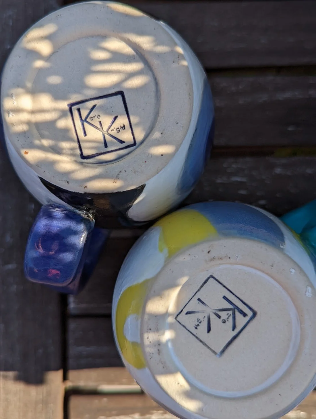Two ceramic mugs with the brand logo on the bottom, placed on a wooden surface. One mug shows a blue handle and colorful design, the other has a white base with yellow and blue accents.