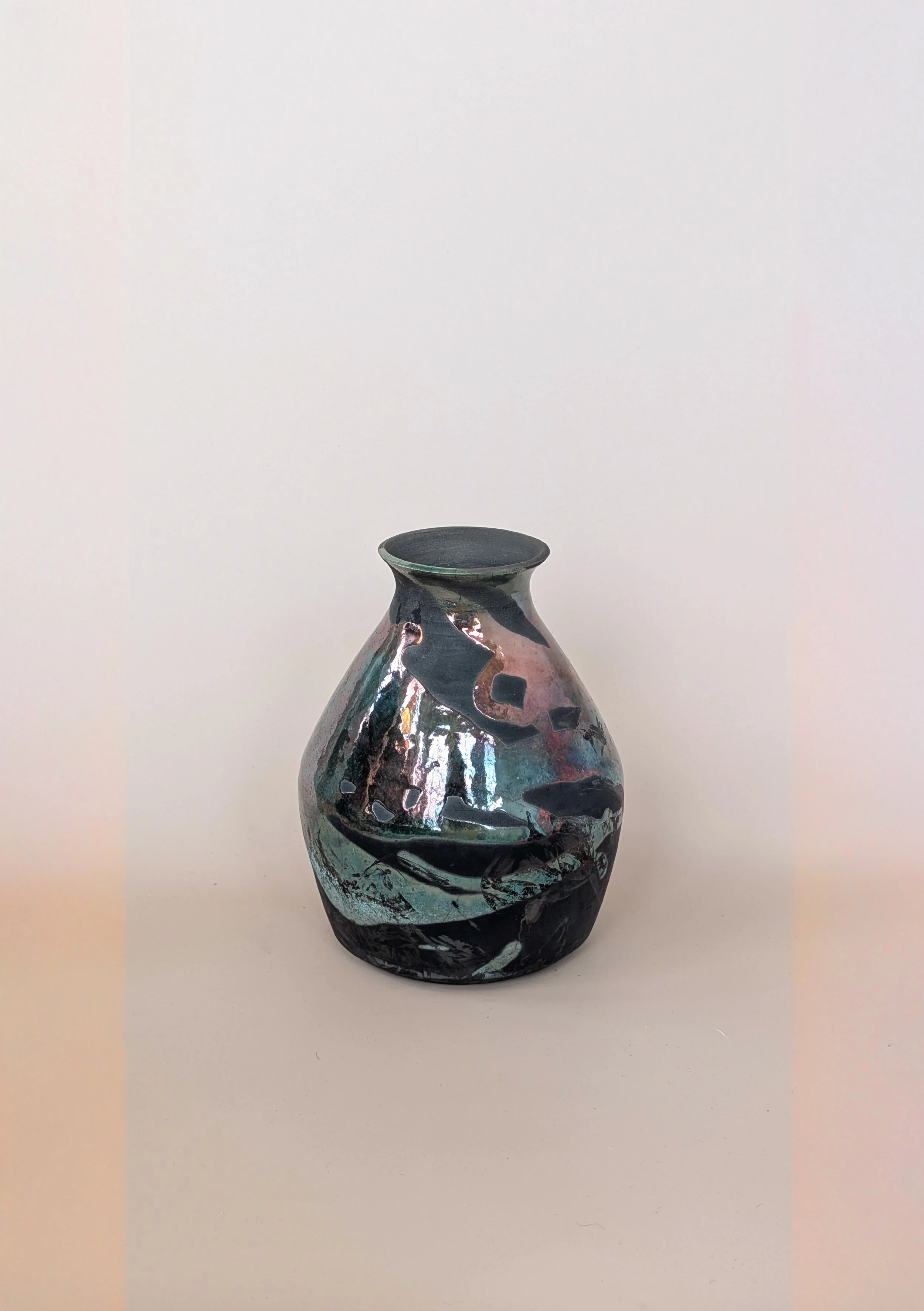 A ceramic vase with abstract, multicolored glaze, featuring shades of black, blue, pink, and iridescent tones, placed on a plain white surface against a white background.