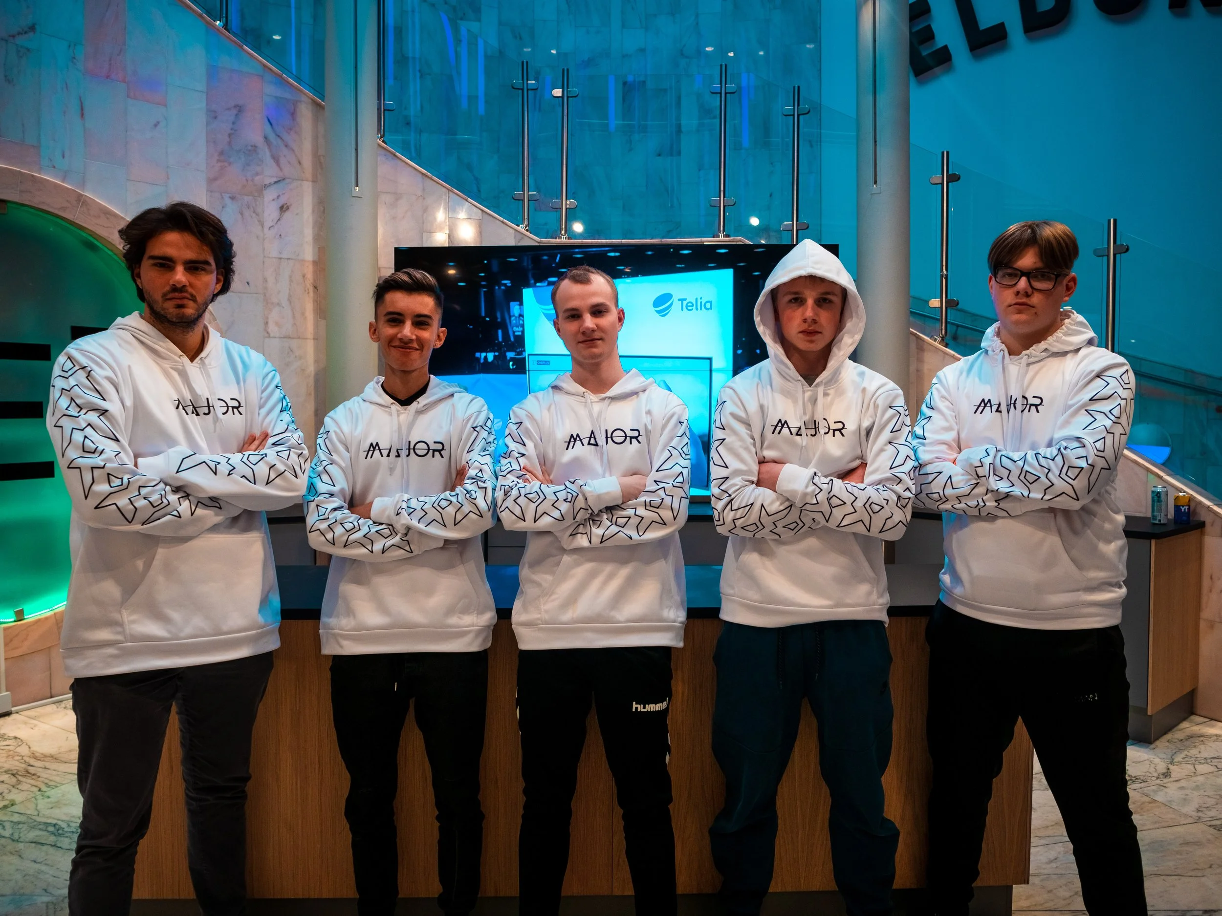 Fem unge menn står med armene i kors foran en bar i en innendørs lobby, iført hvite hettegenser med geometriske mønstre og ordet 'Major' på.