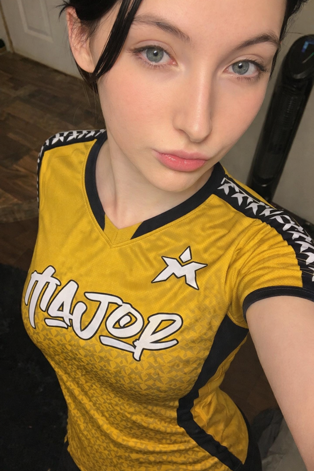 Nærbilde av en kvinne med lett sminke og kort svart hår, iført en gul sportstrøye med svart og hvit detaljering, tatt innendørs.