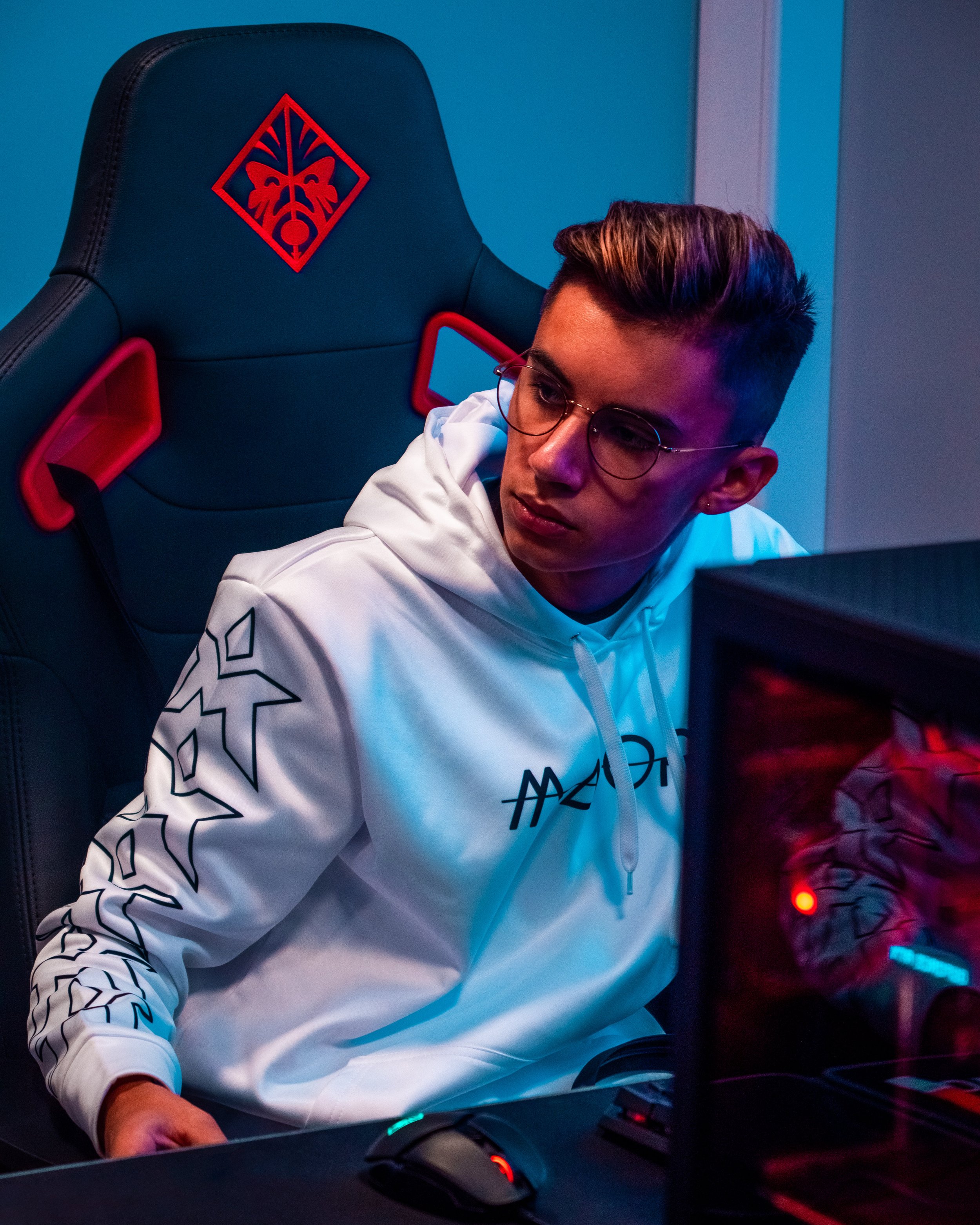 En person med briller i en hvit hettegenser sitter foran en dataskjerm i et gaming-rigg, med en gaming-stol med et H.P. Lovecraft-motiv.