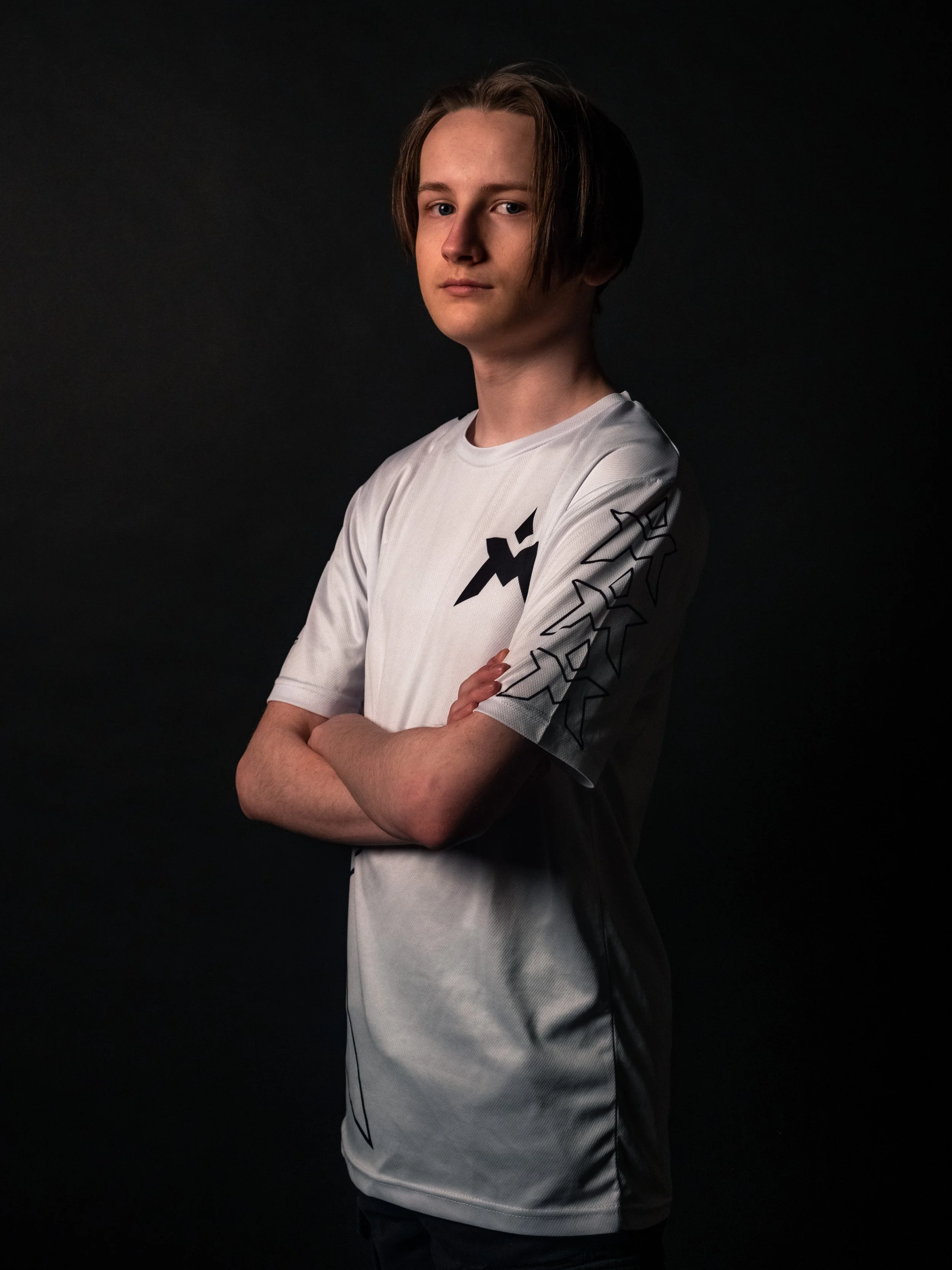 Ung mann med langt hår, kledd i en hvit sports t-skjorte med sorte grafiske design, ser direkte inn i kameraet med armene korslagt, mot en mørk bakgrunn.