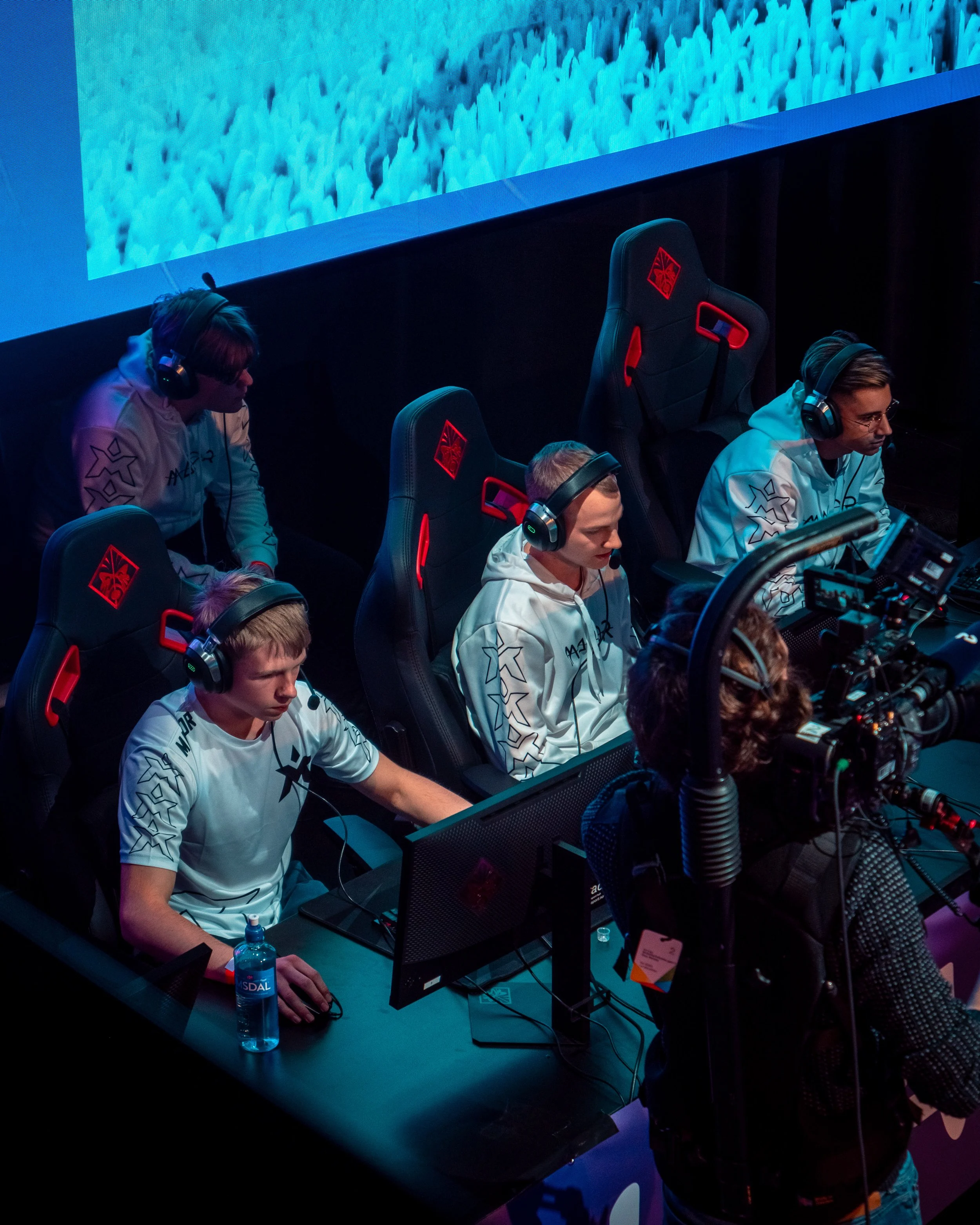 E-sport team som spiller videospill med gamingutstyr, i et mørkt rom med skjermer og profesjonelt kamerautstyr.