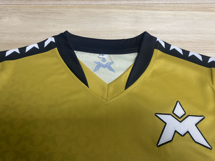 Nærbilde av en gullfarget sports jersey med svart og hvit detaljering rundt halsen og på skuldrene, med en stor logo på brystet.