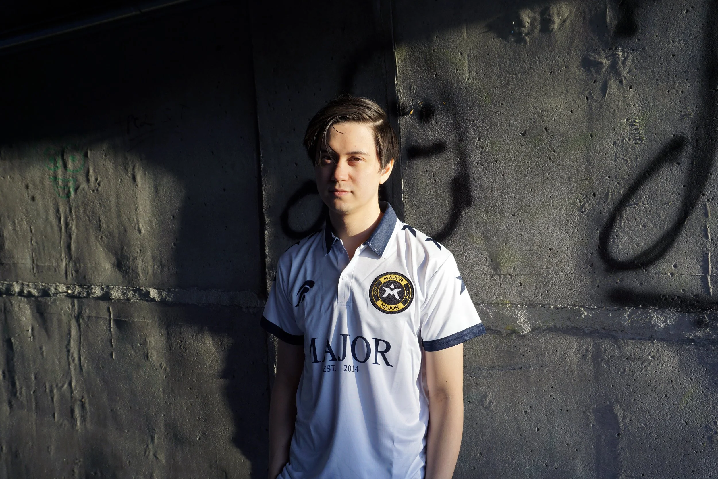 En ung mann med kort, mørk hår, står foran en mørk betongvegg med graffiti, iført en hvit sports T-skjorte med logo og tekst.