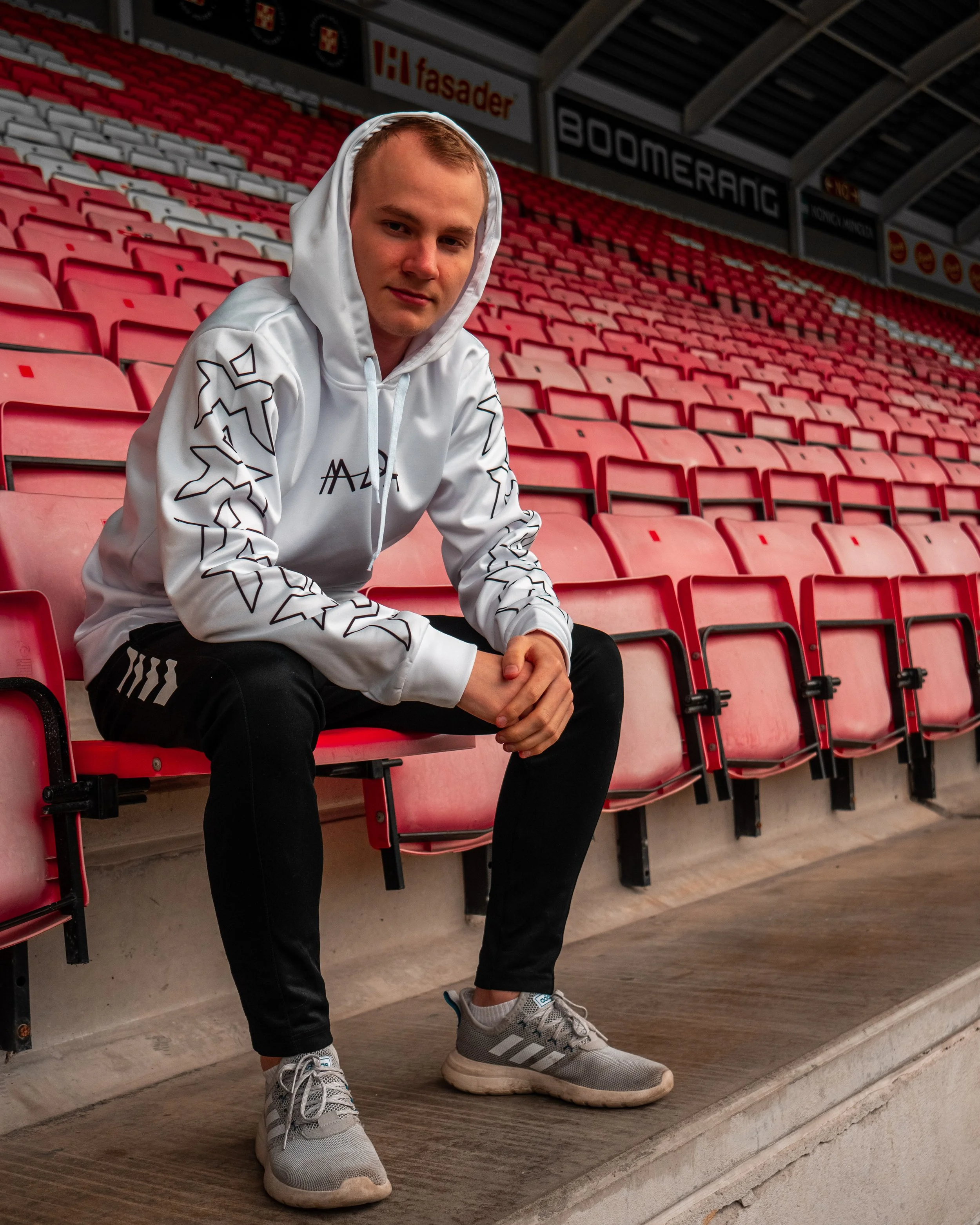 En ung mann i en hvit hettegenser og svarte bukser sitter på rød stadionbenk.