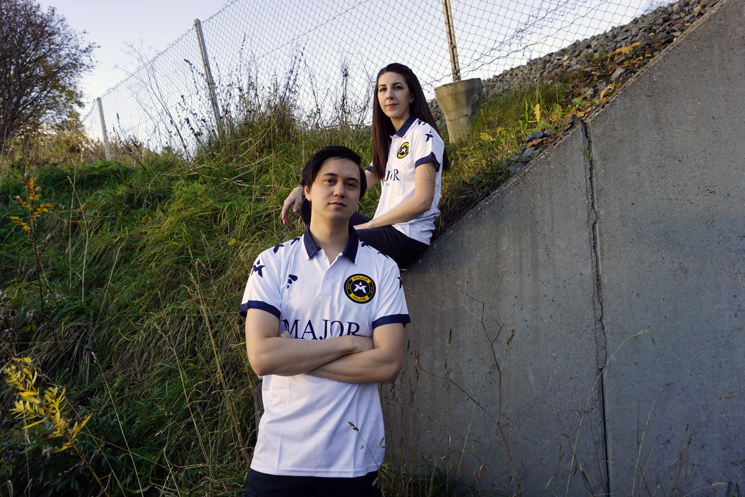 To personer som sitter på en skråning med gress og stein, iført hvite fotballdrakter med logo og tekst, foran en høy gjerde og en steinmur, utendørs.