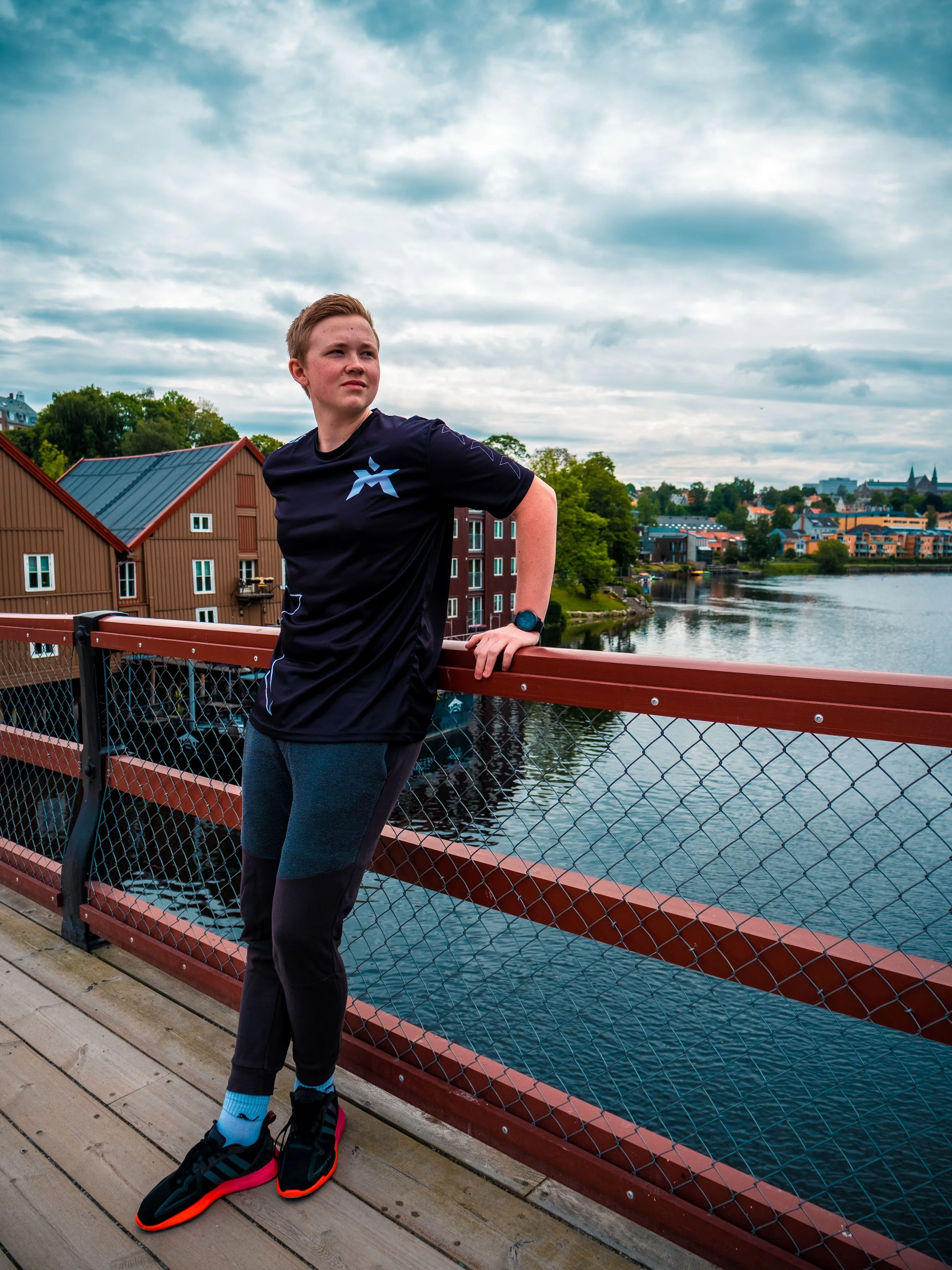 Ung mann står på en trebro ved en elv, iført svart treningstøy, løpesko og armbåndsur, med by og tre i bakgrunnen og skyet himmel.
