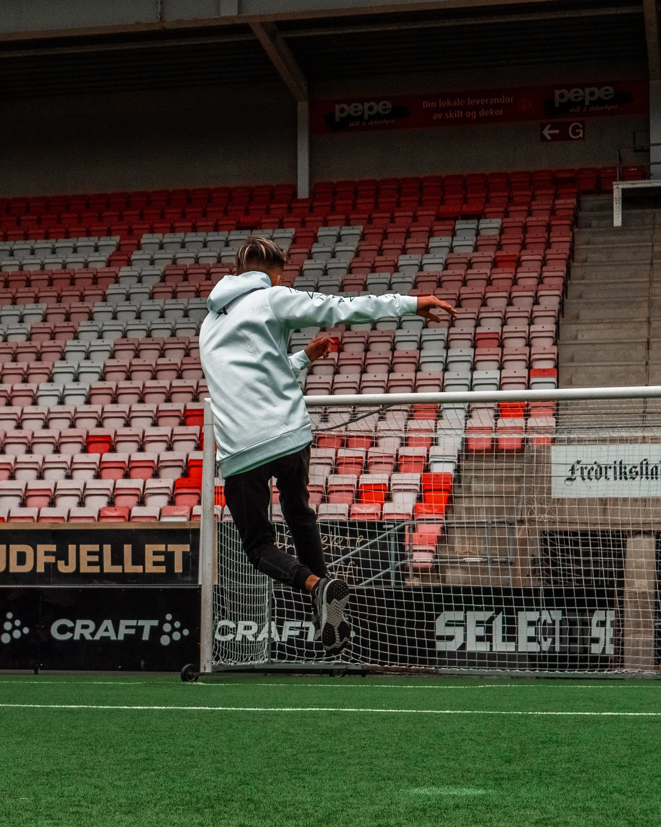 Person i lys grå hettegenser som hopper over fotballmålet i en fotballstadion.