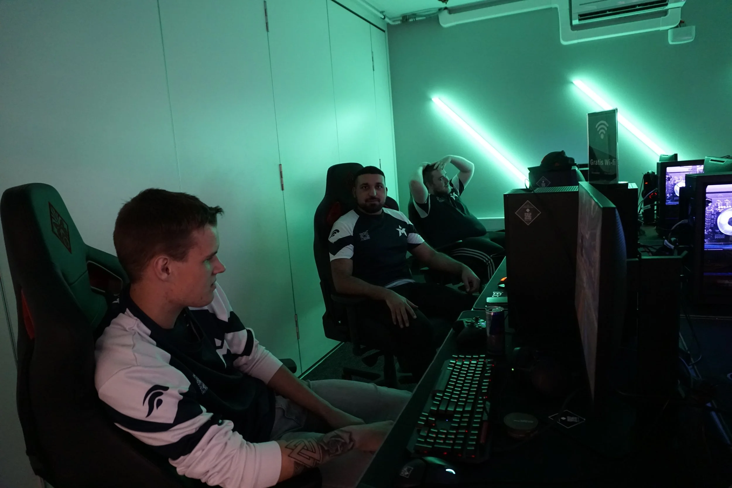 Tre personer som spiller dataspill i et rom med grønne neonlys, to med hodetelefoner, to sitter foran dataskjermer, en person ser mot kamera.