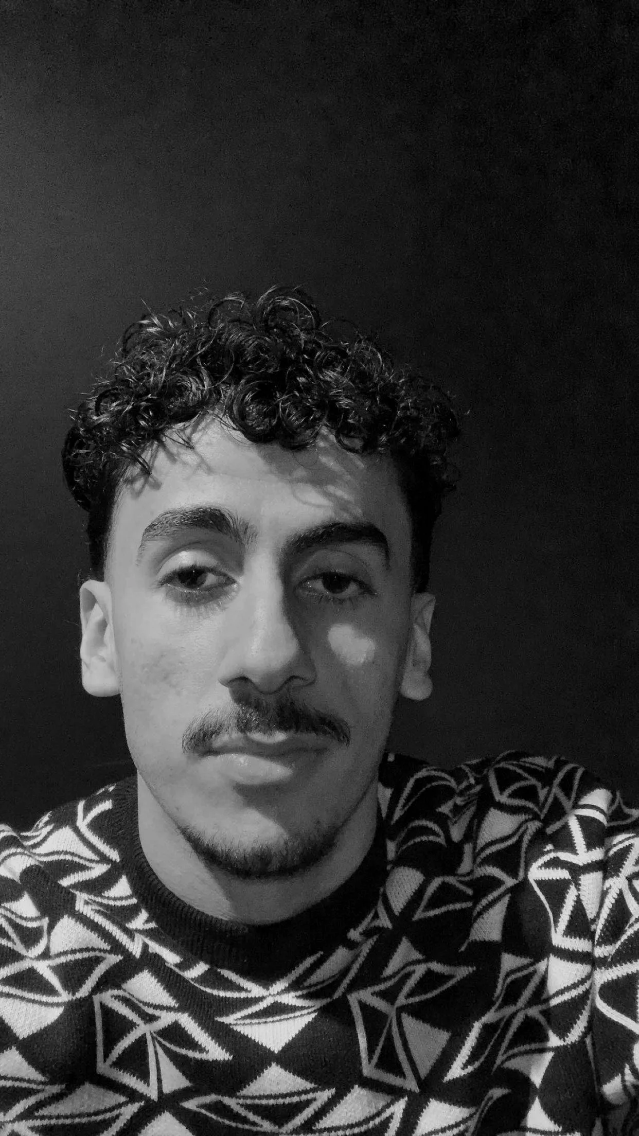 Sort-hvitt portrett av en ung mann med krøllete hår, små skjegg, og en mønstrete genser.