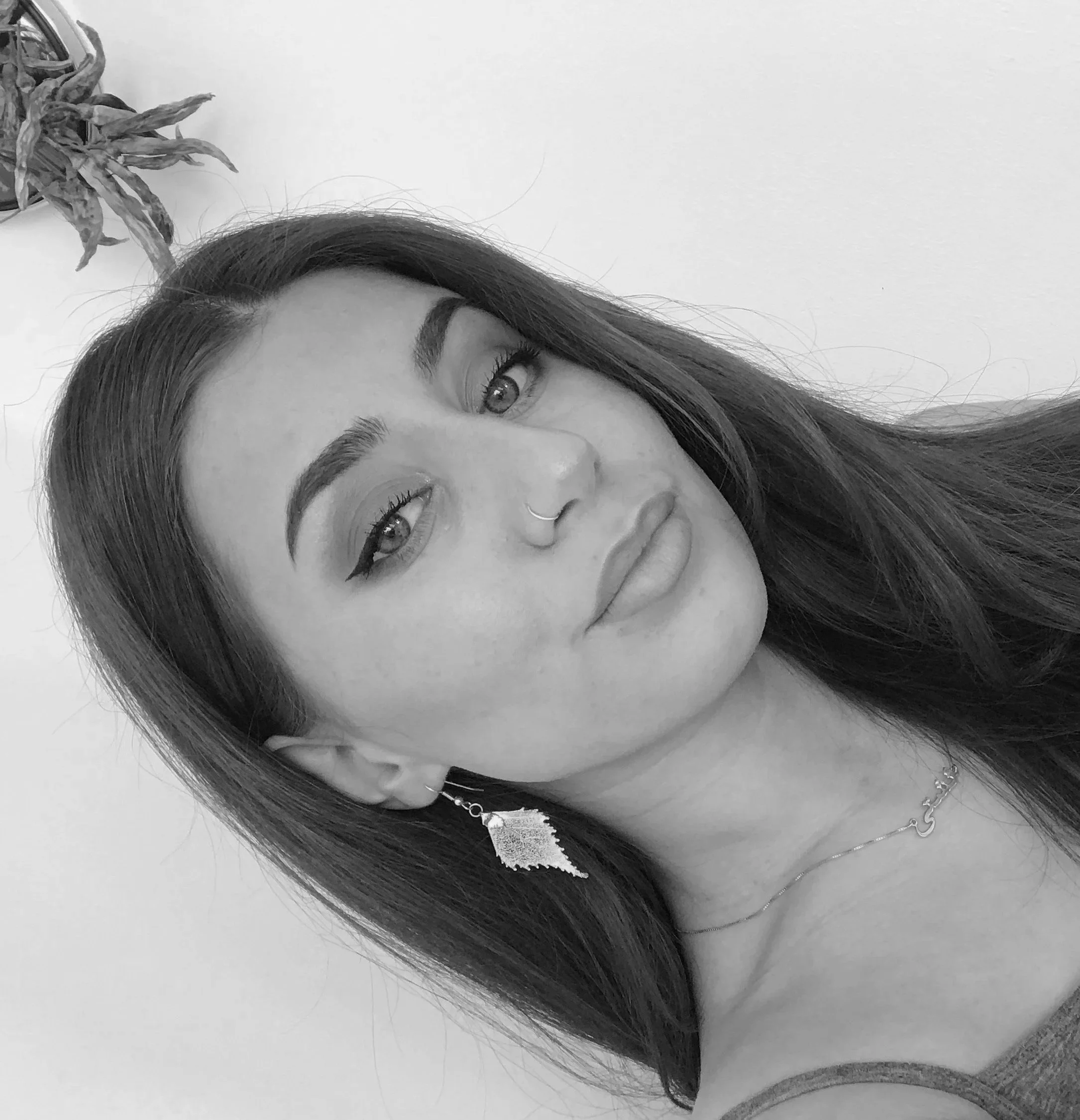 Svart-hvitt portrett av en kvinne med langt mørkt hår, med ring i nesen, øyenbryn med makeup, og øyesminke. Hun har en ørering i øret og en tynn kjede rundt halsen. Bakgrunn med en plante i hjørnet.