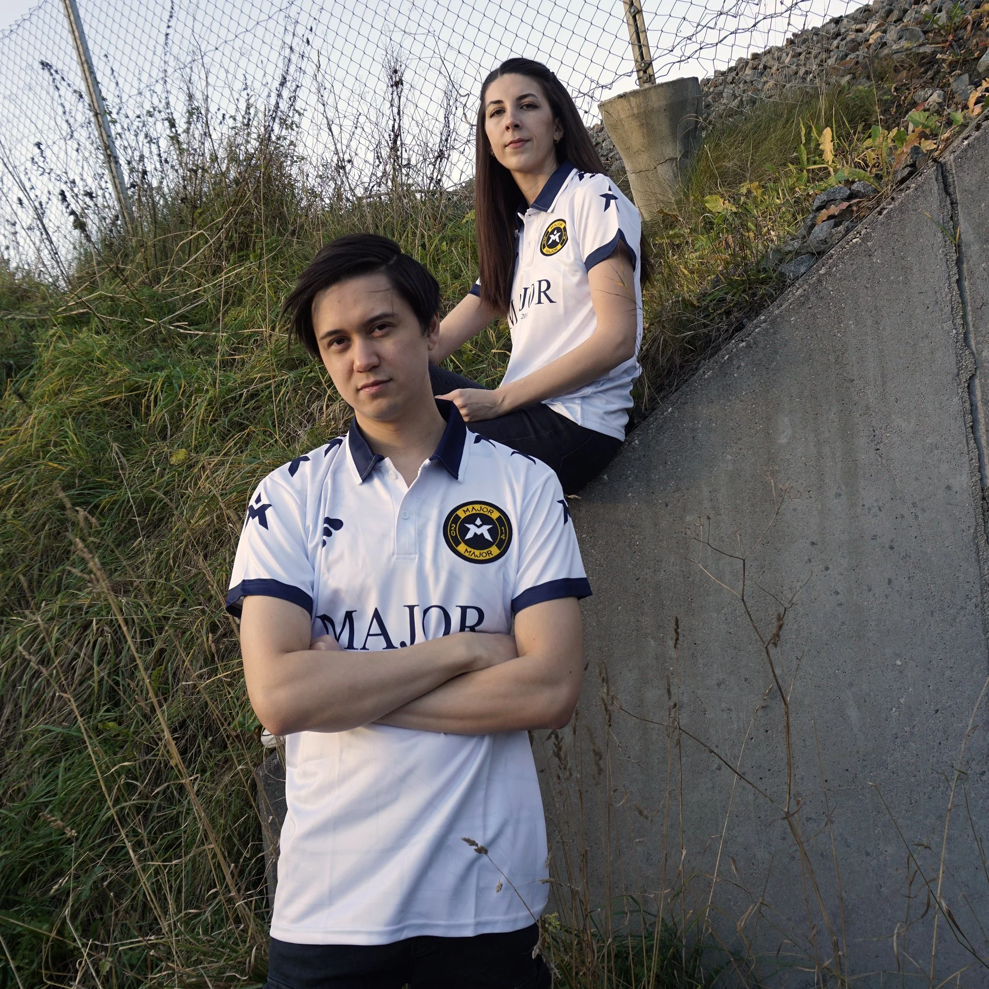 To unge personer kledd i hvite sportsdrakter med logoer, utendørs med gress og trådnett i bakgrunnen.