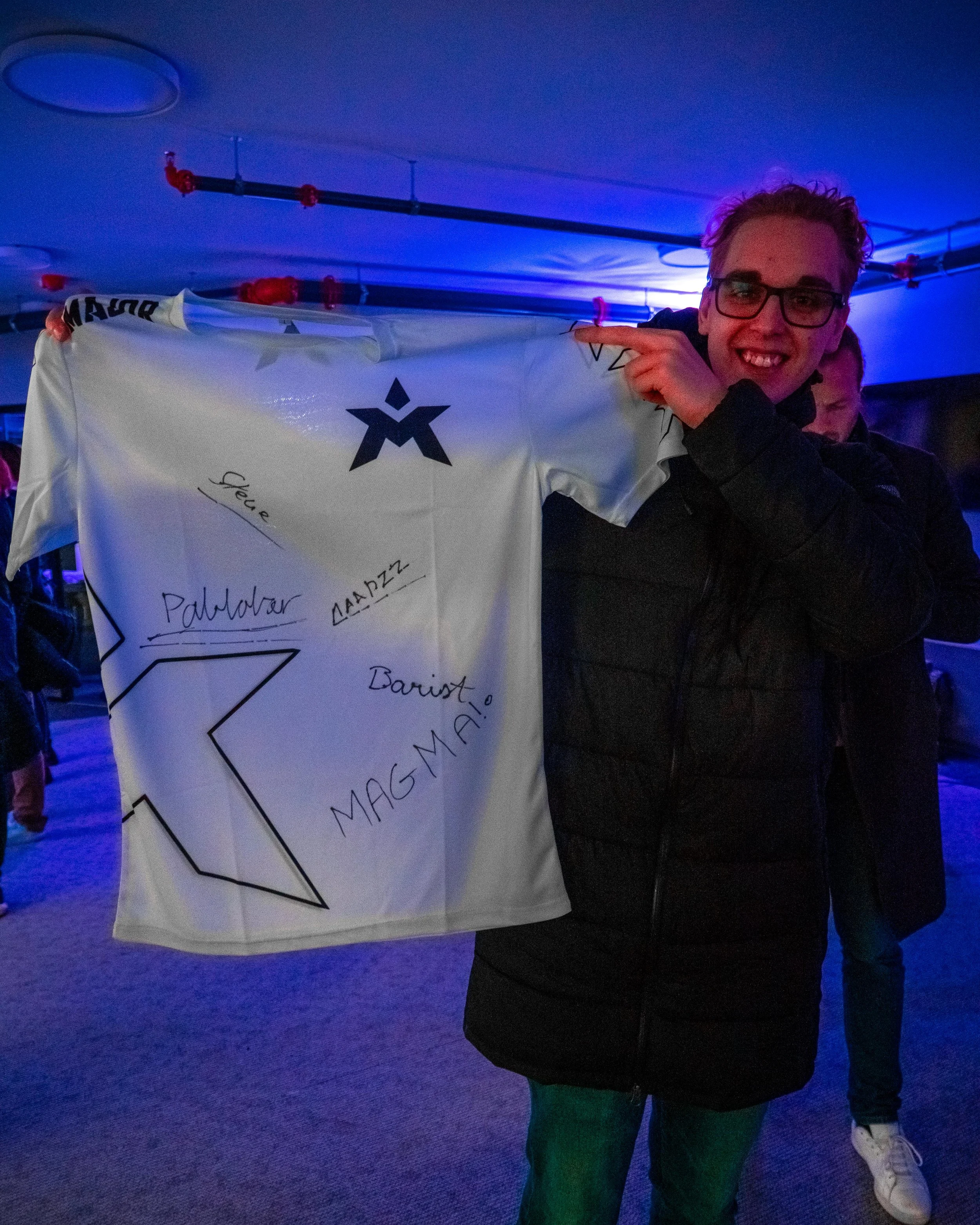 En person smilende holder opp en signert matchtrøye på et arrangement med blå belysning.